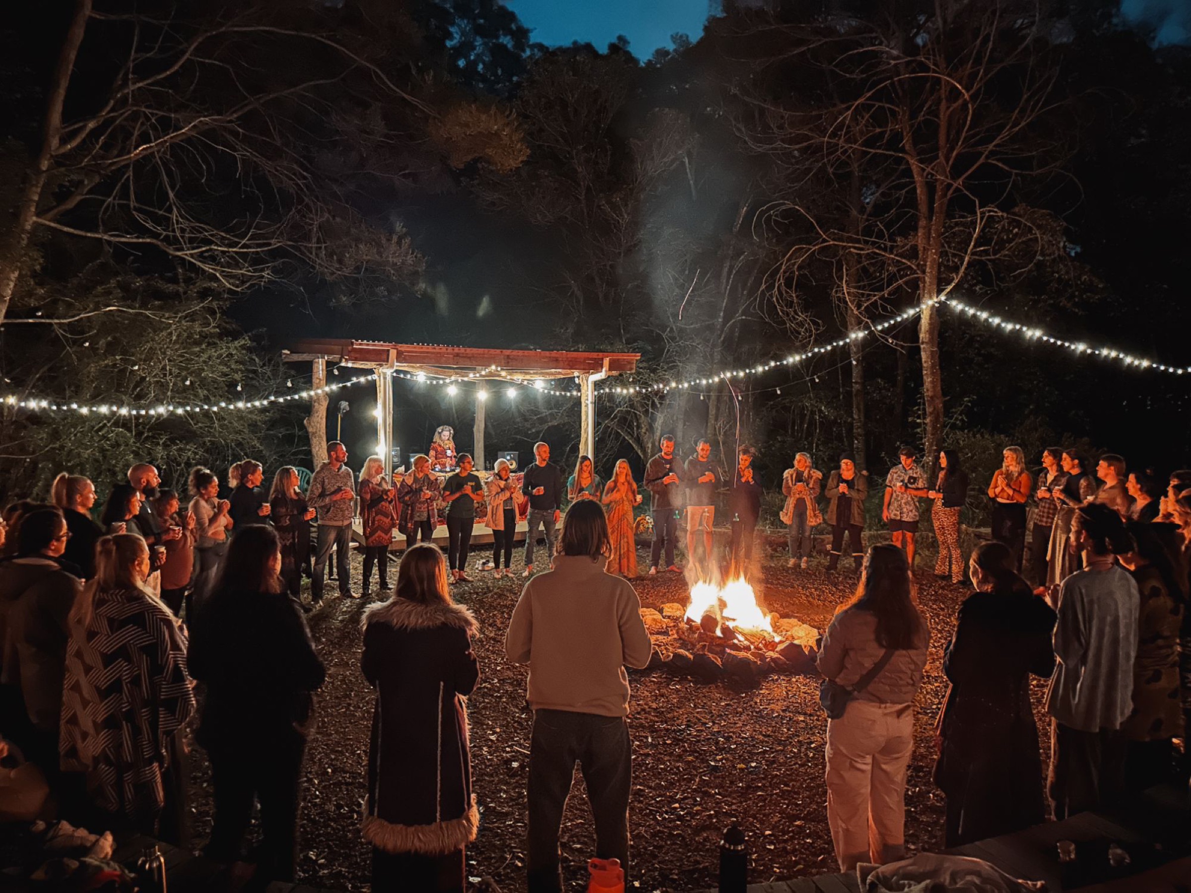 A large circle gathered around a fire at night beneath festoon lights in the bush