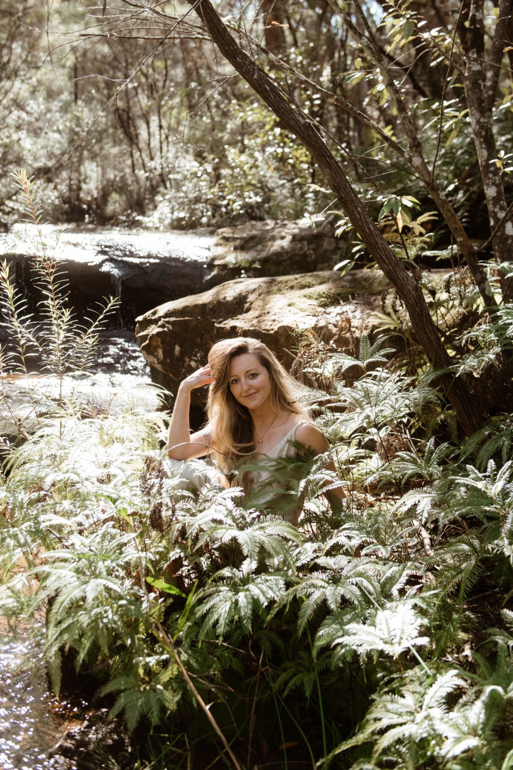 Louisa Fern sitting among ferns beside a bush creek, smiling