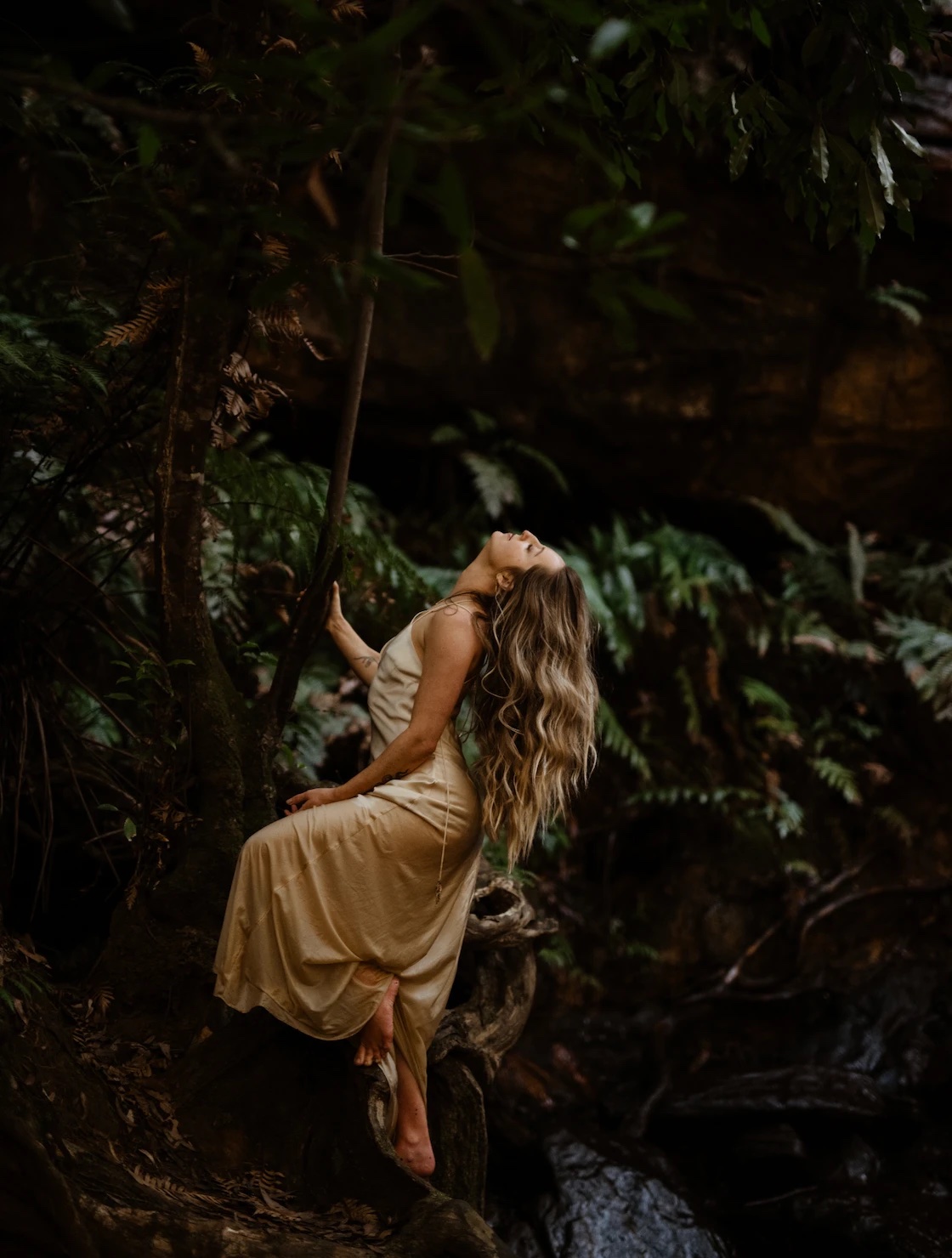 Louisa leaning back against a tree among ferns, eyes closed, in a golden slip dress
