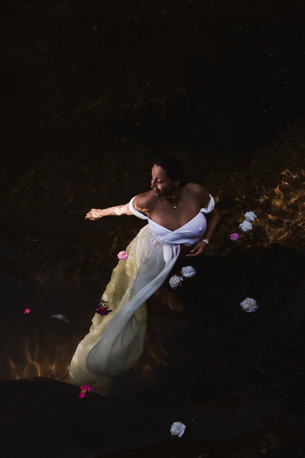 A woman in a flowing white dress standing in dark water scattered with white flowers, photographed from above